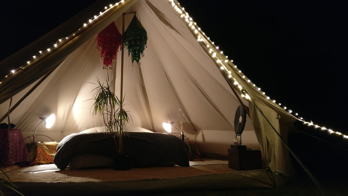 The interior of a well-lit tent showcases a spacious bed with comfortable bedding. Soft lamps and decorative lanterns provide gentle illumination, while a small fan and a potted plant enhance the atmosphere. Decorative textiles and a natural floor cover contribute to a cozy environment.