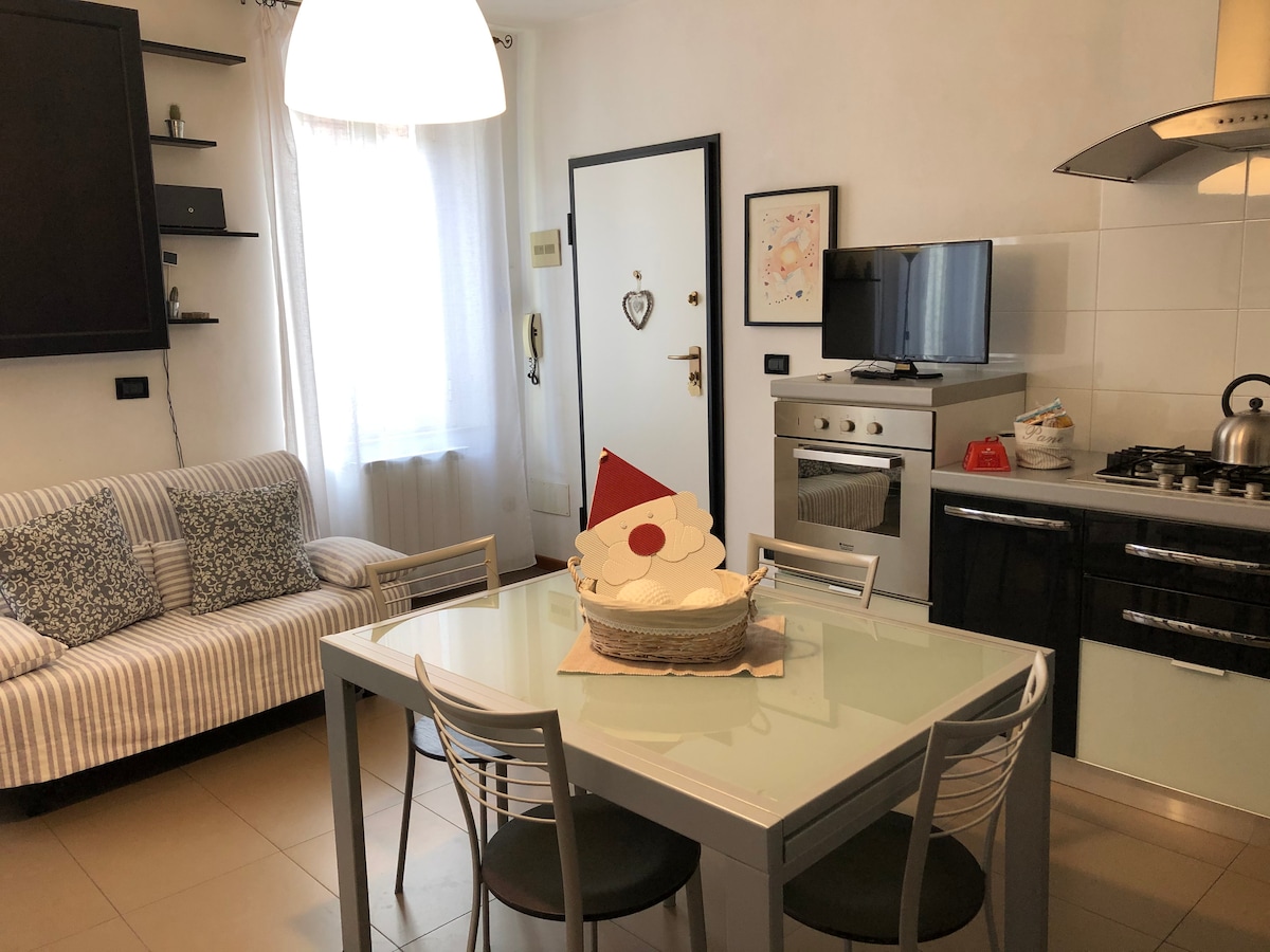 A modern kitchen area is highlighted by a glass dining table with four metal chairs. A light fixture hangs above, illuminating the space. A comfortable sofa covered with a striped pattern is positioned against the wall. Kitchen appliances, including a stove and refrigerator, are visible.