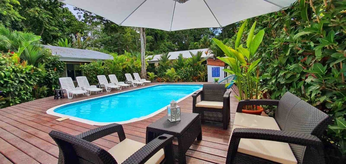 A refreshing outdoor pool is surrounded by a wooden deck, with lounge chairs positioned nearby. Lush green plants line the area, adding a tropical feel. A shaded seating arrangement includes rattan furniture, inviting relaxation in the serene environment.