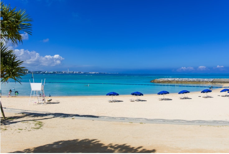 A sandy beach stretches under a clear blue sky, with several blue umbrellas providing shade on the shore. Calm waters meet the beach, and a lifeguard stand is positioned near the water. In the distance, a coastal pathway and a rocky jetty are visible.
