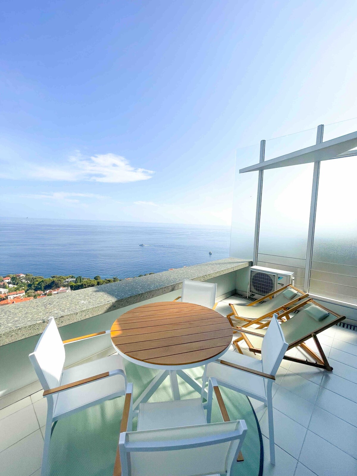 An outdoor terrace features a round wooden table surrounded by four white chairs. A wooden lounge chair is positioned nearby, offering a resting spot. The backdrop presents an expansive view of the sea and the horizon, under a clear blue sky.