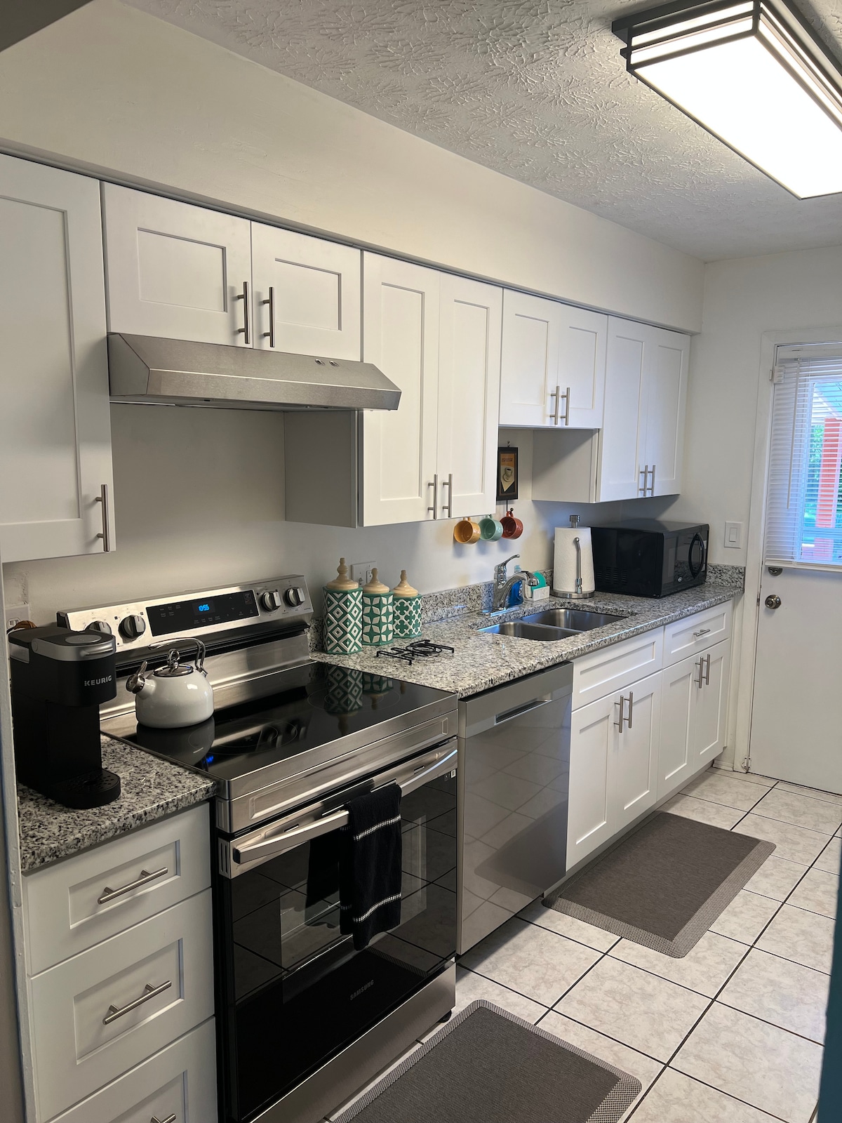 The kitchen features modern white cabinetry and granite countertops, equipped with stainless steel appliances, including an oven and microwave. A coffee maker and various kitchen tools are positioned along the counters. A door leads to an outdoor space, allowing natural light to enter.