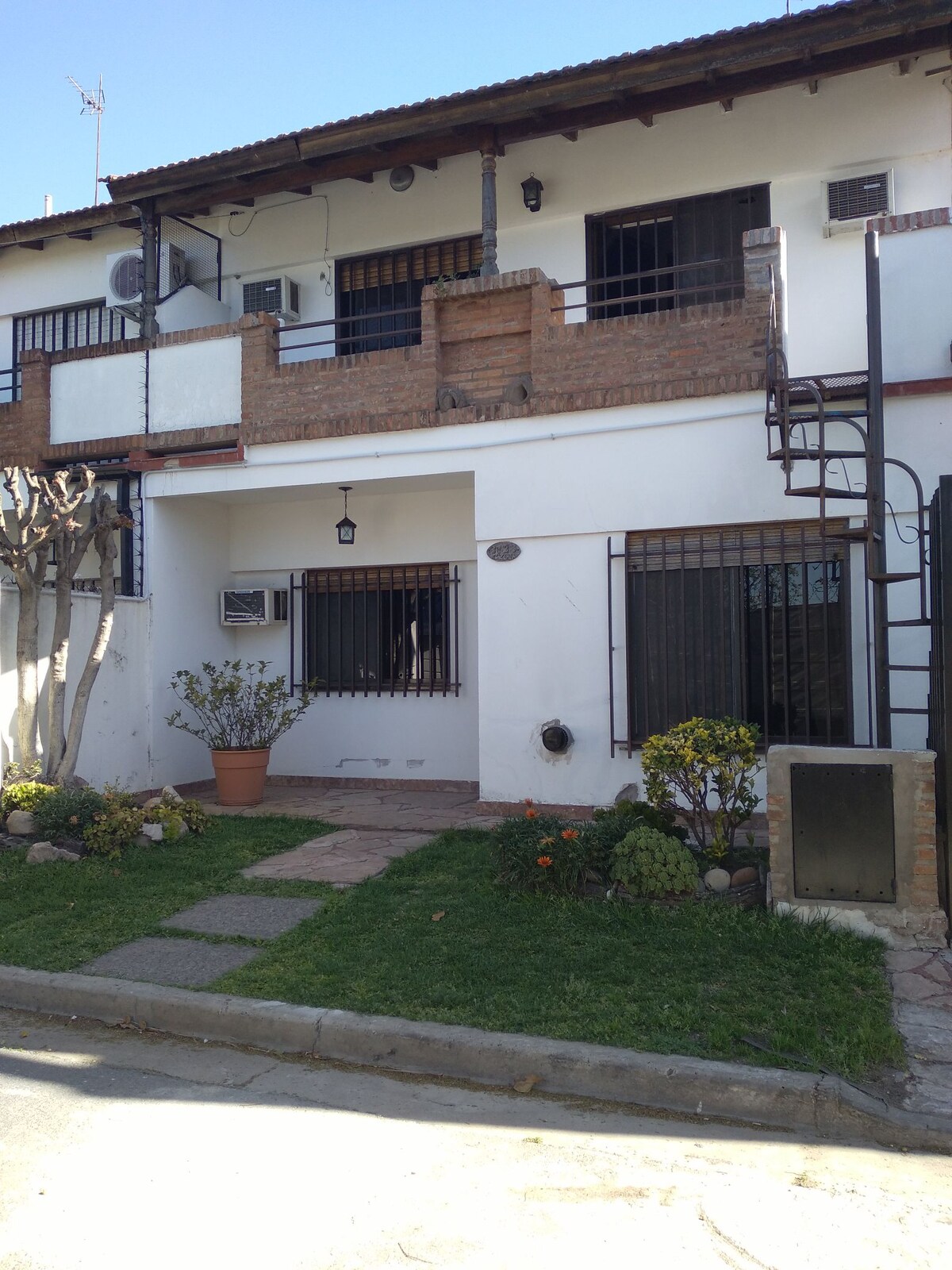 The exterior of a two-story building displays a combination of brick and white walls, featuring a small porch area with potted plants. Windows and balconies provide a glimpse of natural light. A stone path leads to the entrance, bordered by neatly maintained grass and flower beds.