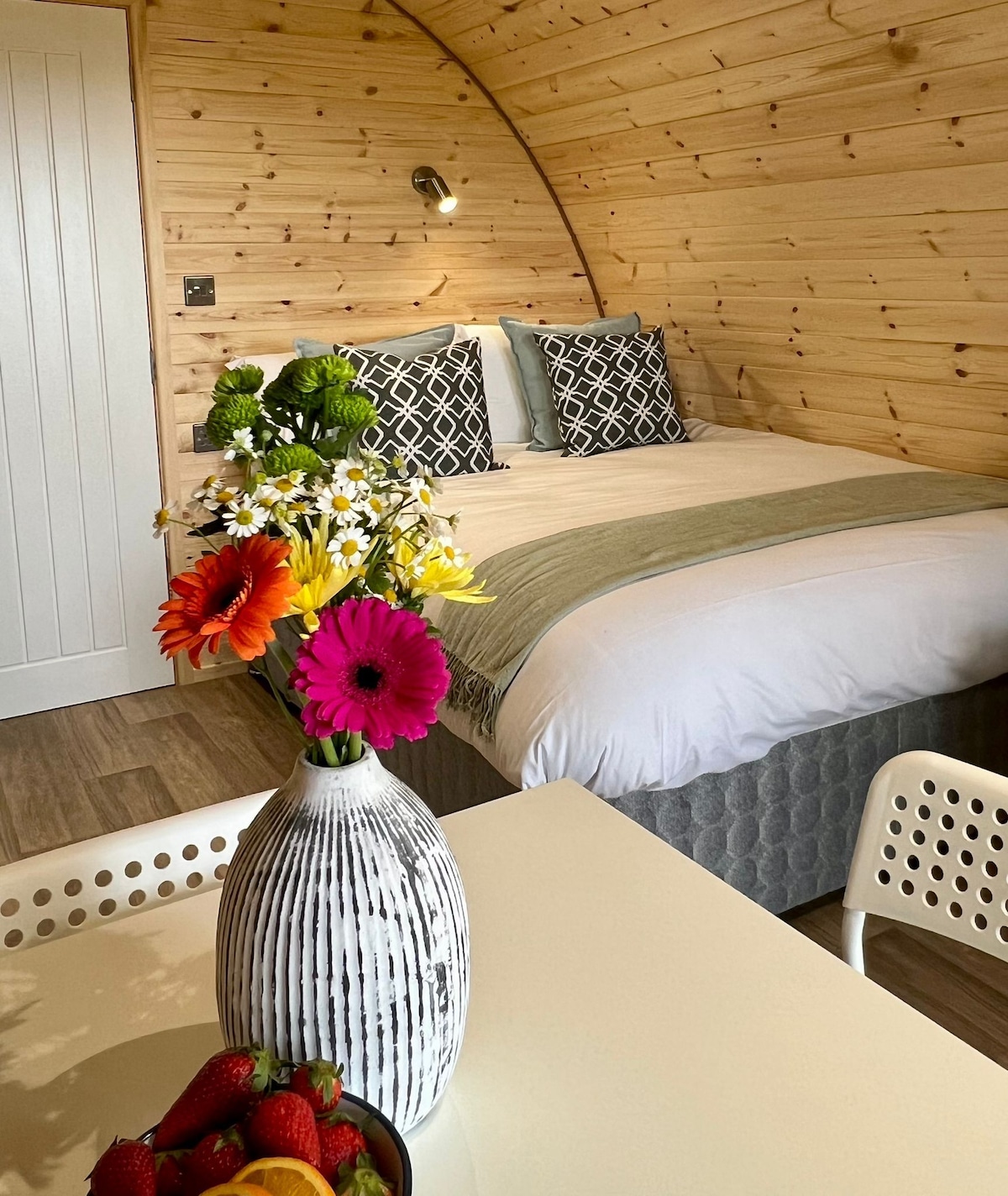 A cozy sleeping area is highlighted by a spacious bed with patterned cushions. A table with a bowl of fresh fruit sits in front of a striking flower arrangement in a vase, set against warm wooden walls that add to the inviting ambiance.