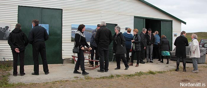 A gathering is taking place outside a barn with a metal exterior. Individuals are seen interacting in a line, with some engaged in conversation. A wooden sled is positioned in the foreground. The landscape is marked by open space and a cloudy sky.