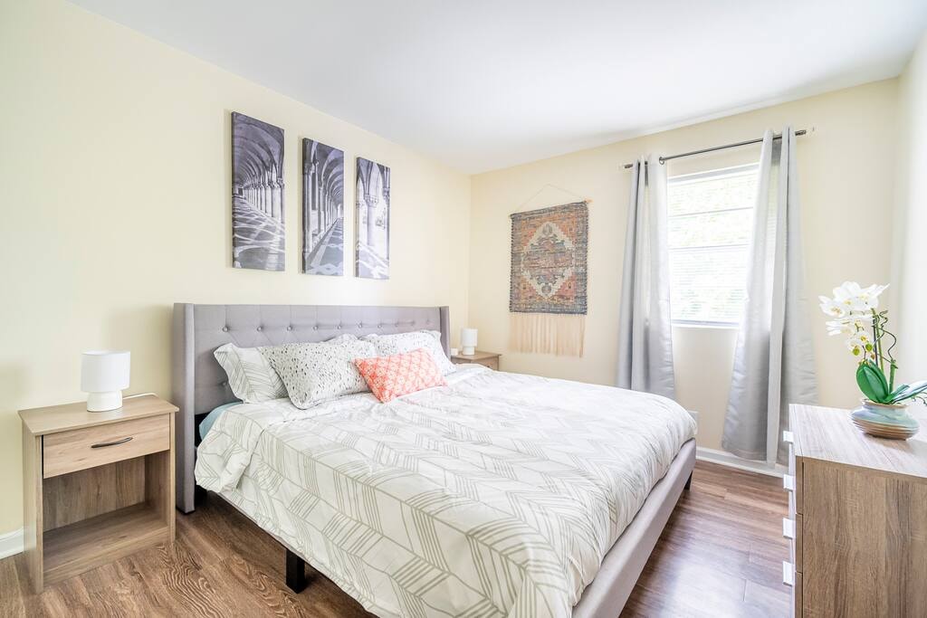 A bright bedroom features a spacious bed with a patterned duvet and decorative pillows. Flanking the bed are two bedside tables with lamps. Wall art above the bed complements the light-colored walls, while curtains frame the window allowing natural light to enter the room.