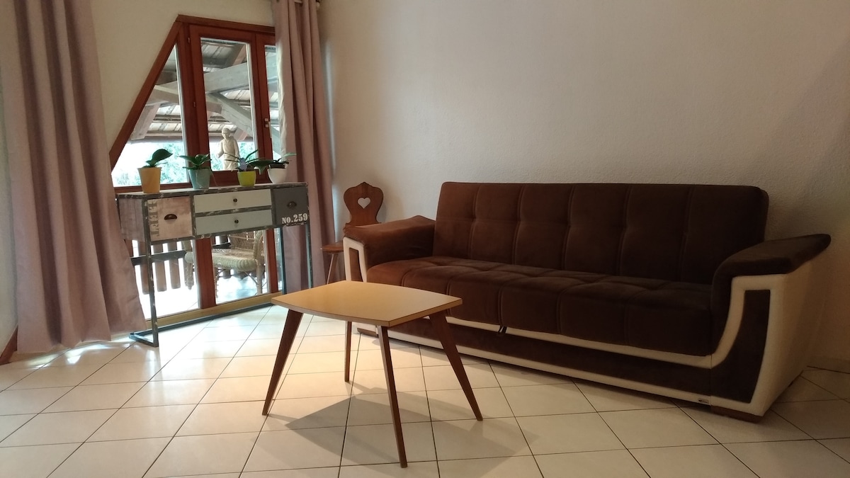 A cozy living area features a brown sofa with a sleek design and wooden legs, accompanied by a light wooden coffee table. Natural light enters through large glass doors that lead to a balcony, framed by soft, neutral curtains.
