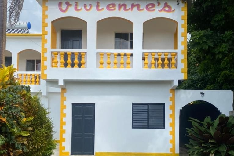 The exterior of Vivienne's showcases a welcoming two-story structure with a bright yellow and white color scheme. Decorative balustrades adorn the balconies, while a dark door and windows add contrast. Lush greenery surrounds the entrance, enhancing the inviting atmosphere.