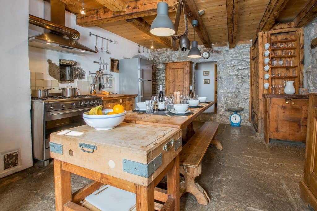 The kitchen features a rustic wooden dining table set for a meal, with decorative bowls of fruit. Exposed wooden beams and stone walls contribute to the farmhouse aesthetic. Modern cooking appliances are positioned along the back wall, complementing the traditional furniture.