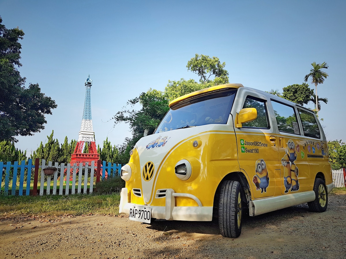A cheerful yellow vehicle with cartoon characters is parked along a tree-lined path. In the background, a replica of the Eiffel Tower stands among green trees, adding an interesting element to the rural scene.