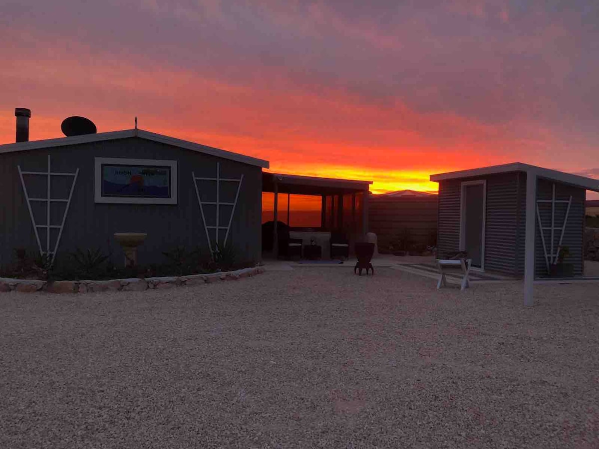 The exterior of the eco cabin is shown at sunset, with vibrant orange and purple skies behind it. The structure features grey siding and large windows, creating a warm and inviting atmosphere. A gravel area surrounds the cabin, and the silhouette of a neighboring outhouse is visible on the right.