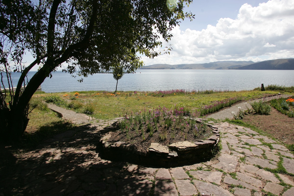 A stone path winds through lush gardens leading to the serene banks of Lake Titicaca. Colorful flowers dot the landscape, and a large tree provides shade, framing the tranquil water and distant hills under a partly cloudy sky.