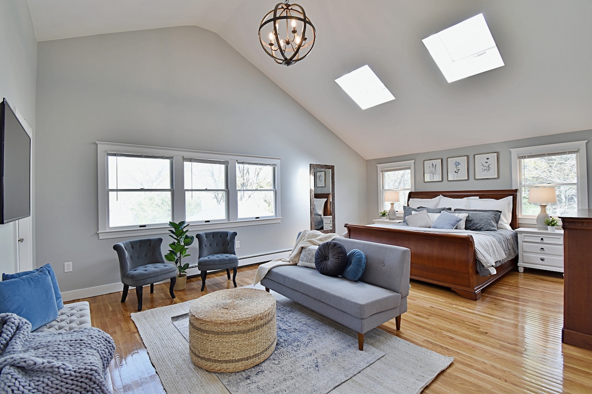A spacious bedroom features a large wooden bed, complemented by gray upholstery on a modern sofa and two accent chairs. Natural light floods the room through multiple windows and skylights, with hardwood floors and a soft area rug enhancing the elegant decor.