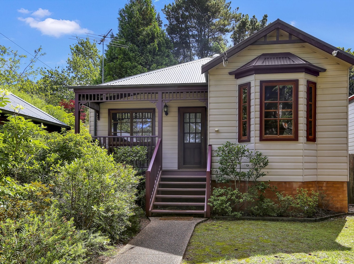 The cottage features a charming exterior with light-colored wood siding and a dark roof. A small front porch is accentuated by a railing and steps leading up to the entrance. Lush greenery surrounds the home, providing a serene, inviting atmosphere.