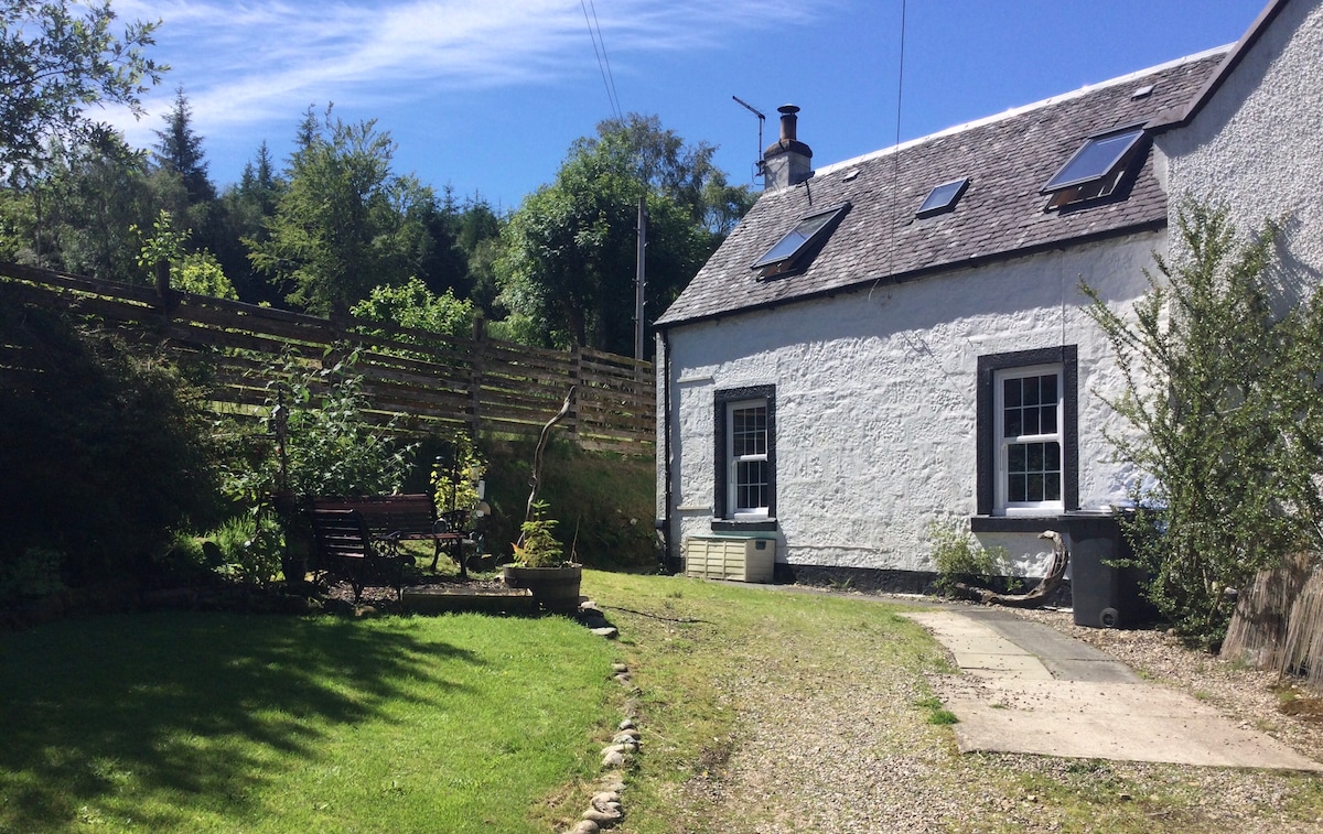 A tranquil outdoor space is featured, showcasing a well-maintained garden area with a lawn and gravel path. The cottage's exterior, painted in white with dark trim, is complemented by seating and a sunny sky above, lending to a peaceful atmosphere.