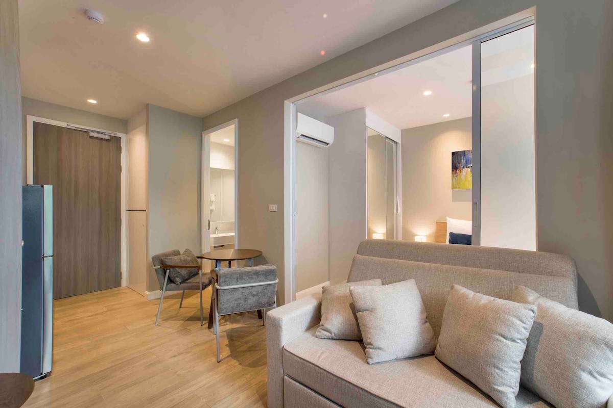 A modern living area is shown, featuring a light-colored sofa with several cushions. A small dining table with two chairs is positioned nearby. The space is brightened by natural light, and a glimpse of a bedroom area is visible in the background through glass doors.