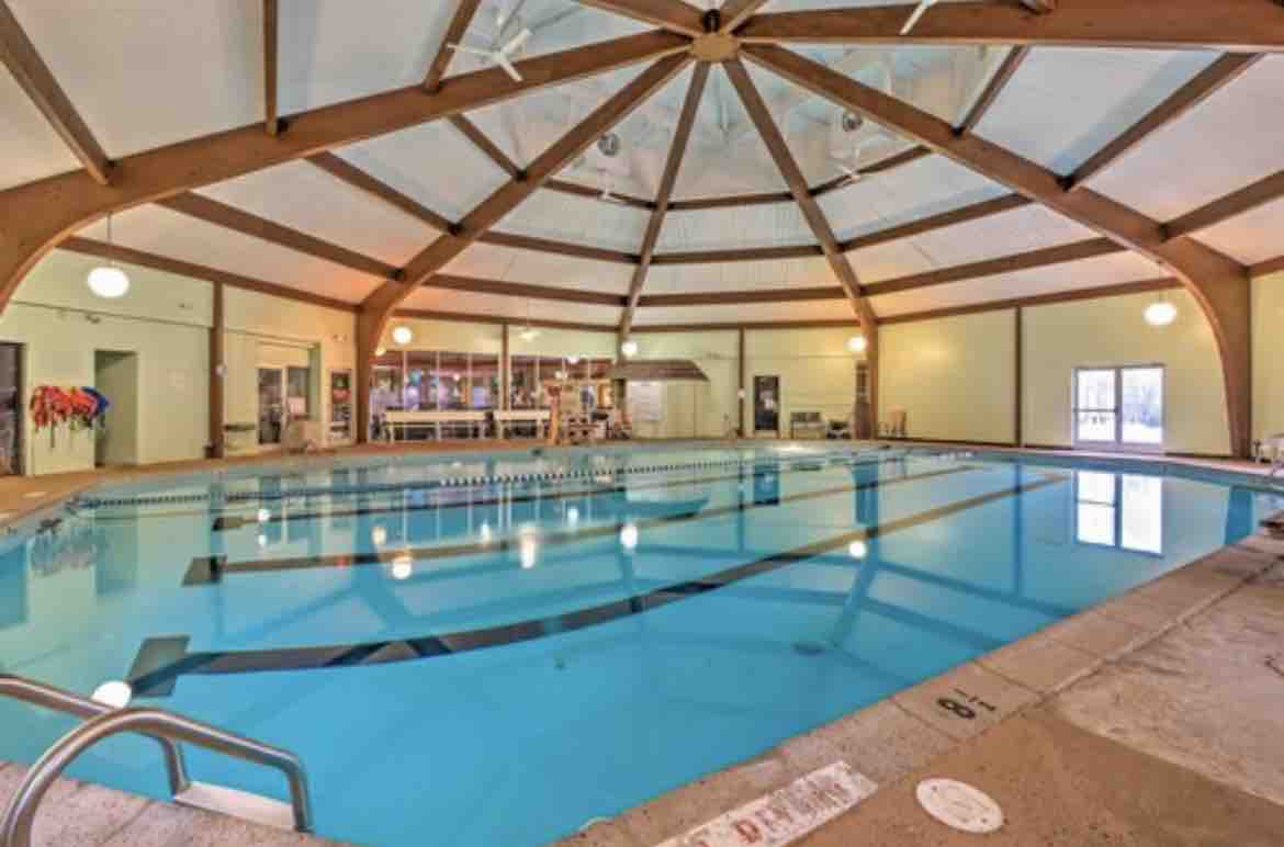 An indoor swimming pool is enclosed within a spacious, dome-like structure. Natural light filters through large windows, illuminating the clear blue water. High wooden beams create an open and airy atmosphere, while seating areas and amenities are visible in the background.