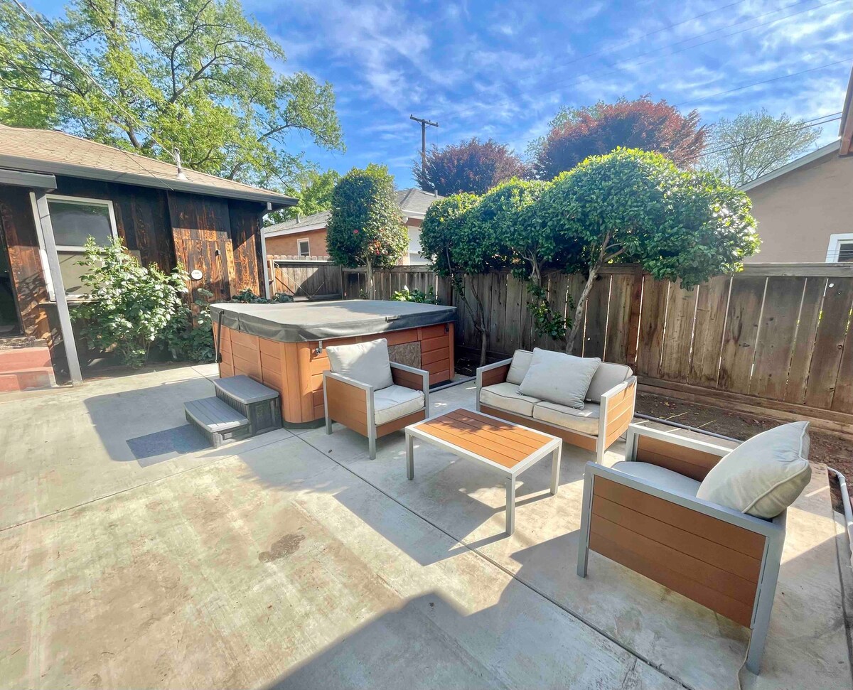 A spacious backyard is enclosed by wooden fencing, featuring a hot tub and a lounge area with light-colored seating and side tables. Potted greenery adds a touch of nature, while the patio surface is made of concrete, creating an inviting outdoor space.