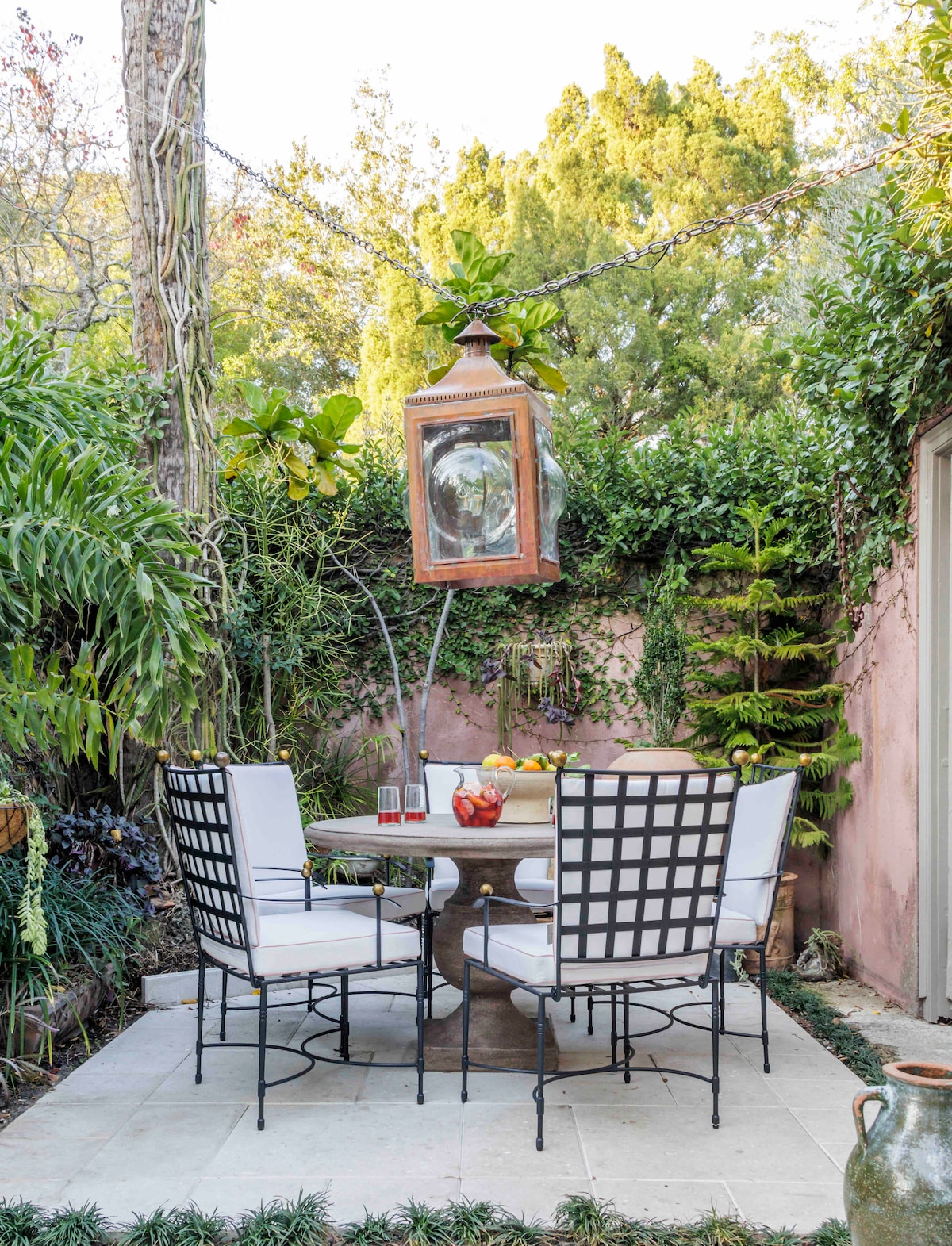 An outdoor dining space features a round table surrounded by four black and white chairs. Lush greenery and plants create a tranquil setting, while a large lantern hangs above, providing ambient lighting. A light-colored stone floor complements the serene atmosphere.