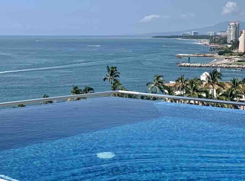 An infinity pool stretches to the horizon, creating a seamless view with the ocean. Palm trees line the landscape, while distant buildings mark the coastline. The sky reflects shades of blue, enhancing the serene atmosphere of the setting.