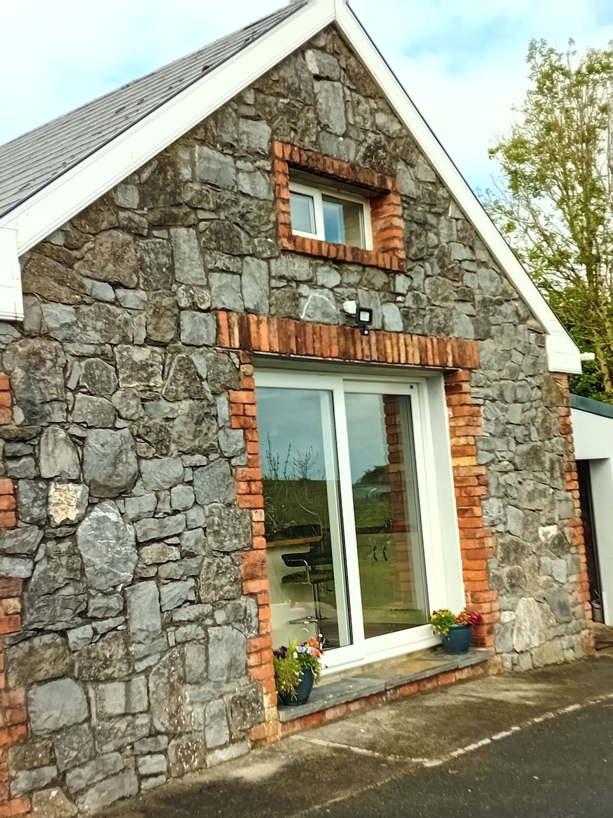 The exterior features a charming stone façade with a combination of grey stone and red brick detailing. Large glass doors provide access to the interior, accentuated by flowerpots on either side, contributing to a welcoming entrance.