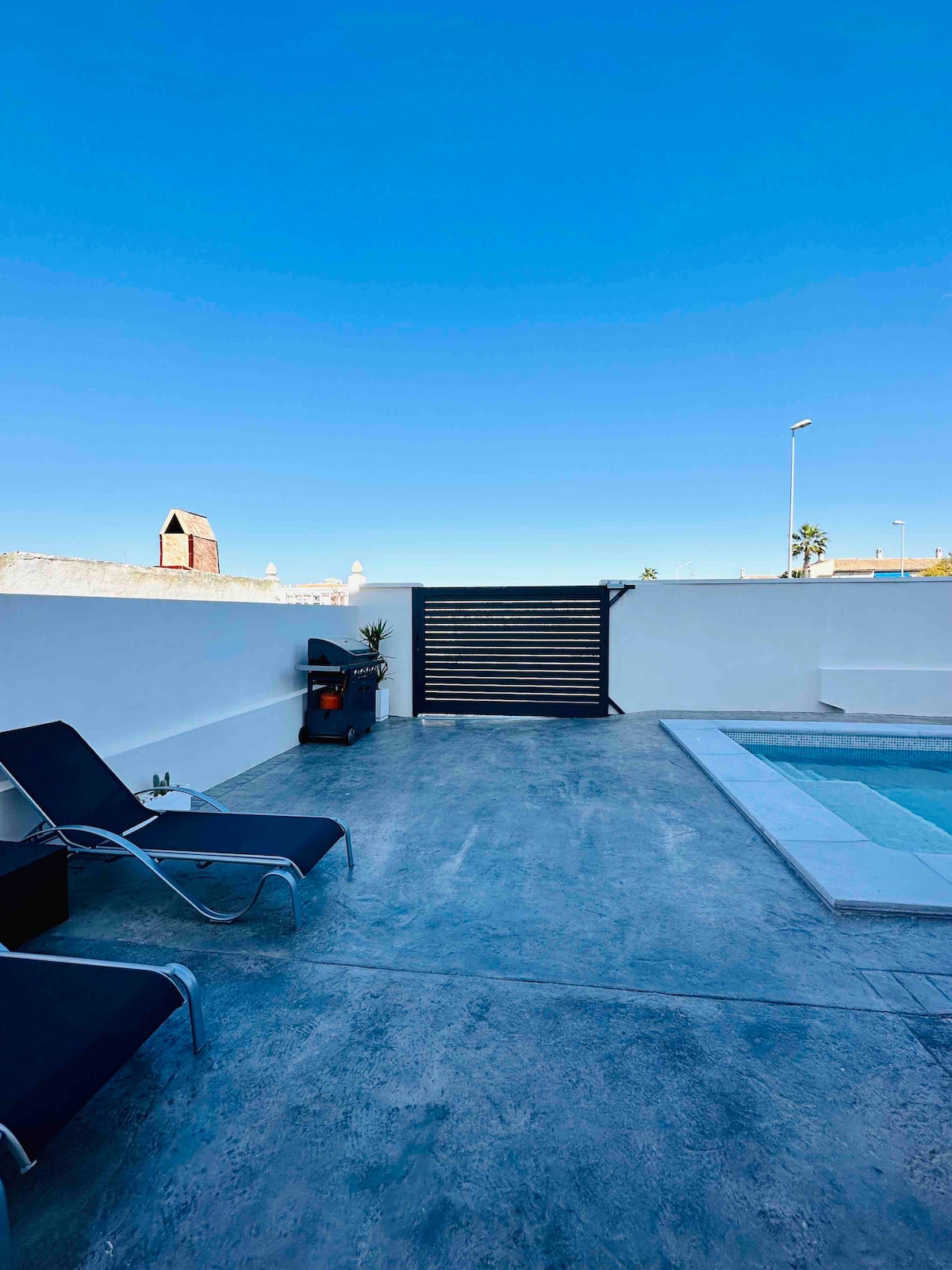 A private terrace is presented, featuring sleek lounge chairs positioned alongside a swimming pool. The space is enclosed with a white wall and a decorative gate, complemented by clear blue skies above. A small plant adds a touch of greenery to the serene outdoor setting.