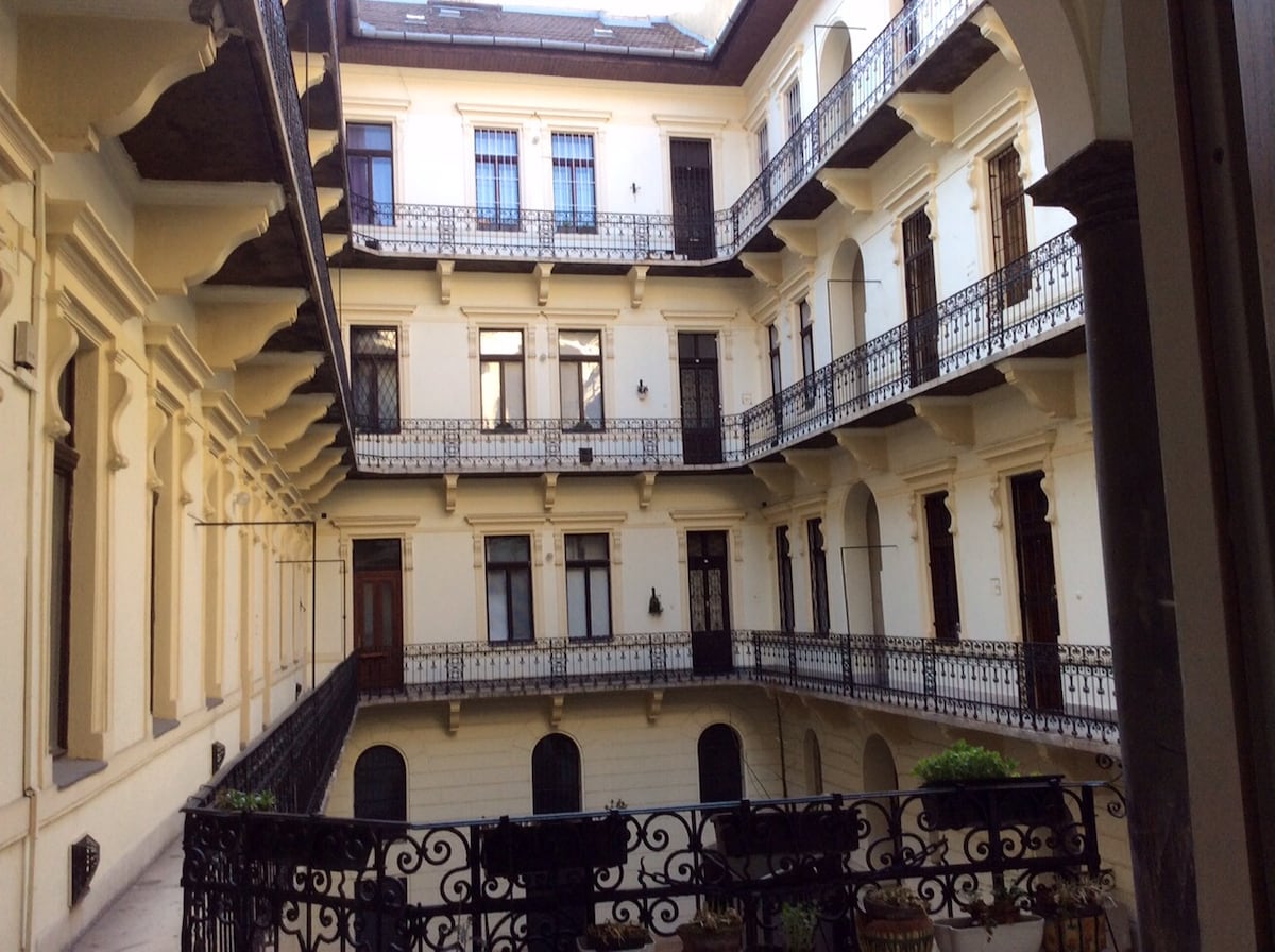 An interior courtyard is framed by elegant arches and wrought iron railings. The light-colored walls of the period building contrast with dark accents. Greenery is visible around the courtyard, offering a serene view of the surrounding architecture.