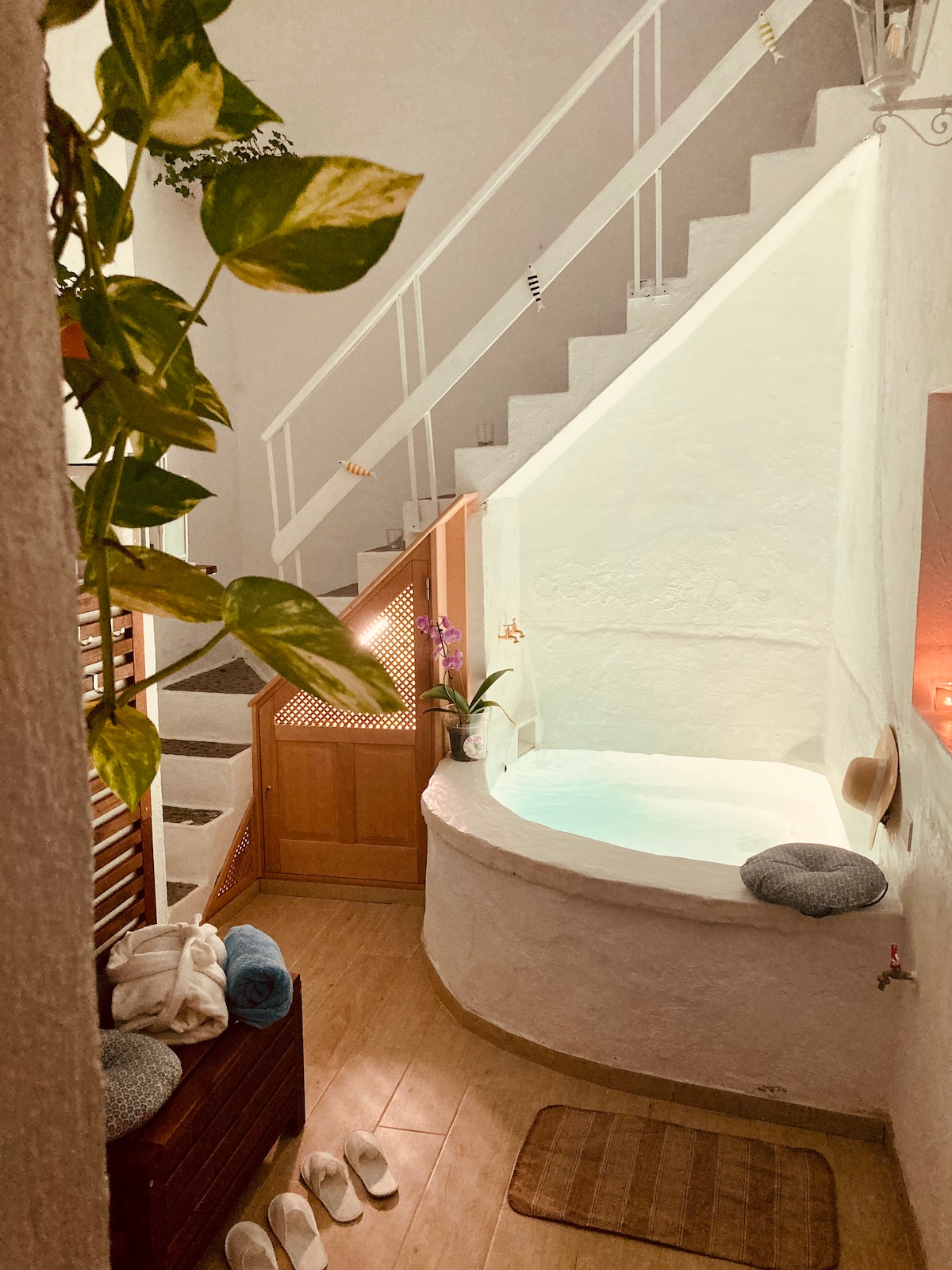 An inviting interior space features a small circular pool with warm ambient water, situated near a staircase. Soft towels and slippers are neatly arranged on a wooden surface, while a plant adds a touch of greenery. Lighting creates a serene atmosphere in this calming nook.