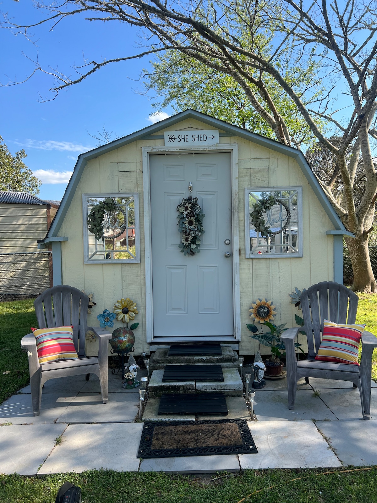 The exterior of the she shed features a charming door adorned with a wreath, flanked by two decorative mirrors. Colorful pillows are placed on the two chairs, and flower decorations add a cheerful touch. A pathway leads to the entrance, surrounded by vibrant greenery.