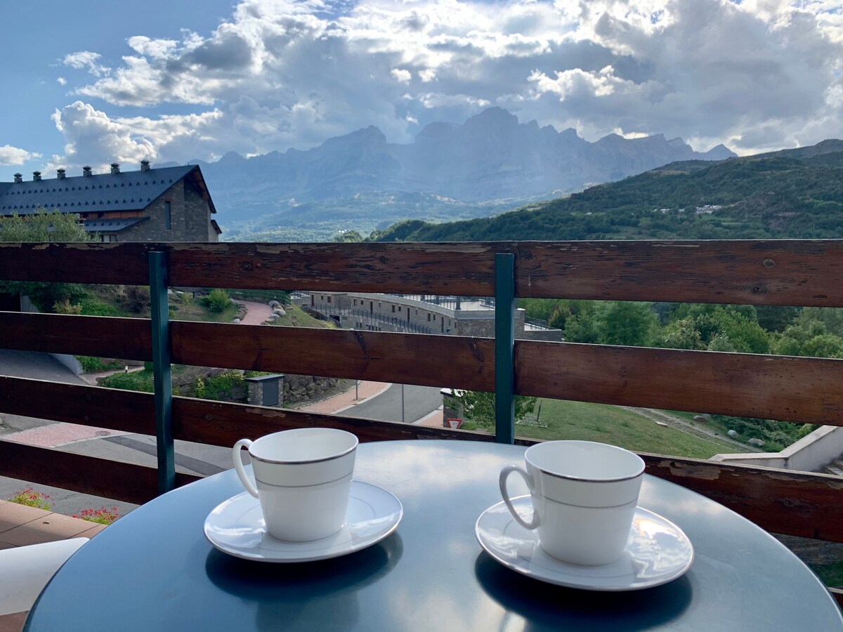 A terrace table is set with two white cups on saucers, overlooking a scenic view of mountains under a partly cloudy sky. The lush green landscape expands below, highlighting the tranquility of the location.