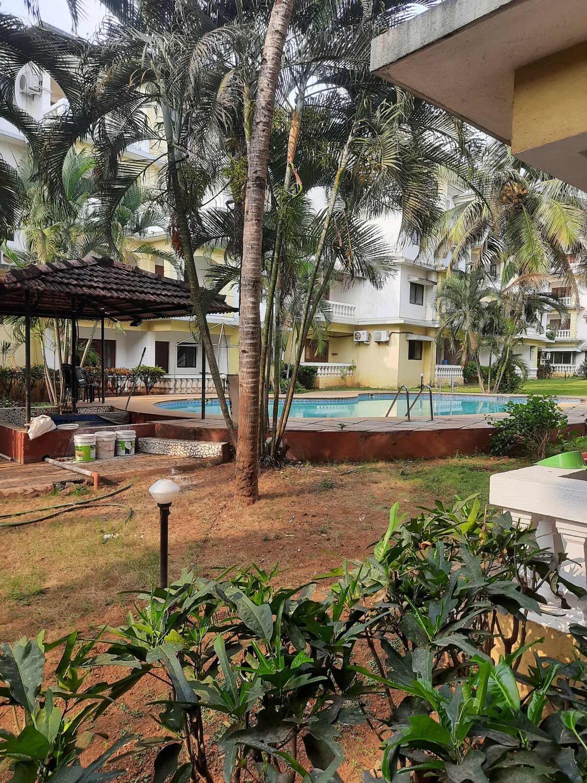 A view of the serene garden area features well-maintained grass and tropical plants. A swimming pool is visible beyond the landscaping, surrounded by palm trees and a shaded seating area. The overall setting provides a peaceful ambiance, ideal for relaxation.