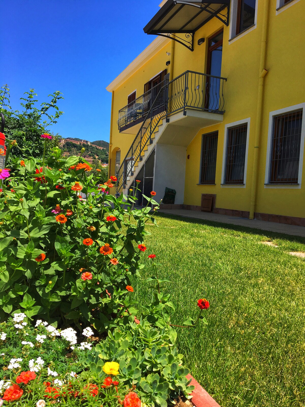 A vibrant garden showcases an array of flowers, including orange and white blooms, against a backdrop of a bright yellow building. An external staircase leads to a balcony, and a well-maintained lawn provides a welcoming green space.