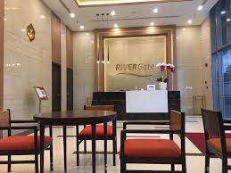 A modern lobby area is presented with sleek, polished floors and elegant lighting. A reception desk is situated at the back, flanked by decorative plants. Several wooden chairs with orange cushions are arranged around a round table, creating a welcoming seating space for guests.