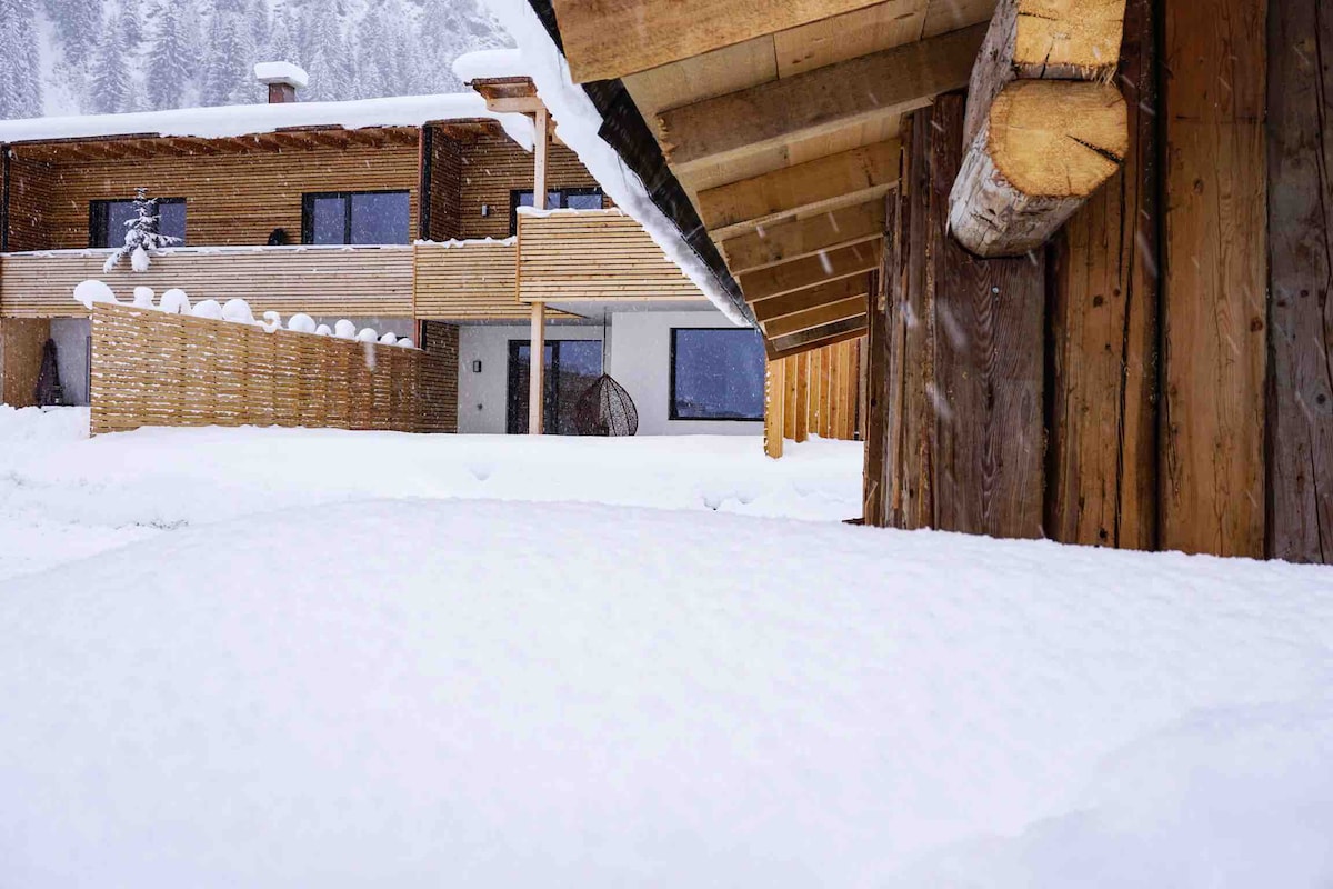 A winter scene captures a wooden structure surrounded by a tranquil blanket of snow. The building features large windows and an inviting entrance, while gentle snowfall adds a serene atmosphere to the landscape.