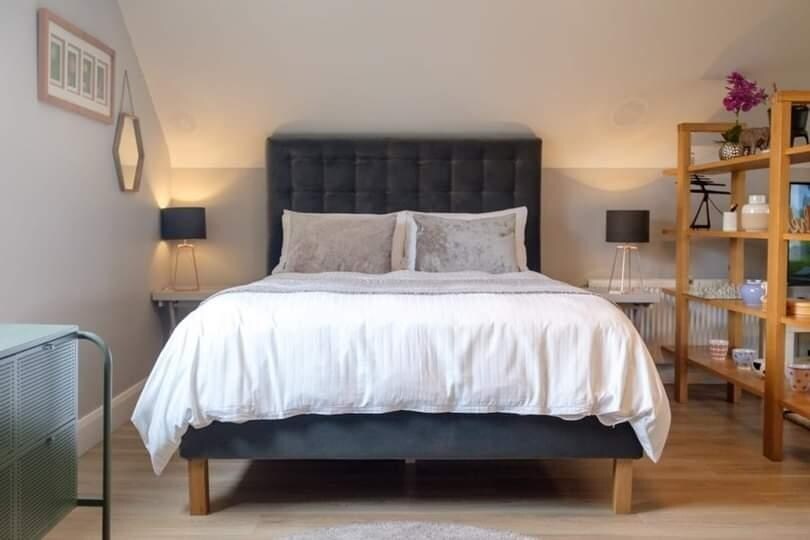 A spacious studio apartment features a soft gray upholstered bed framed by two nightstands with lamps. A wooden shelving unit displays various decorative items and plants. Natural light filters in, complementing the warm tones of the room.