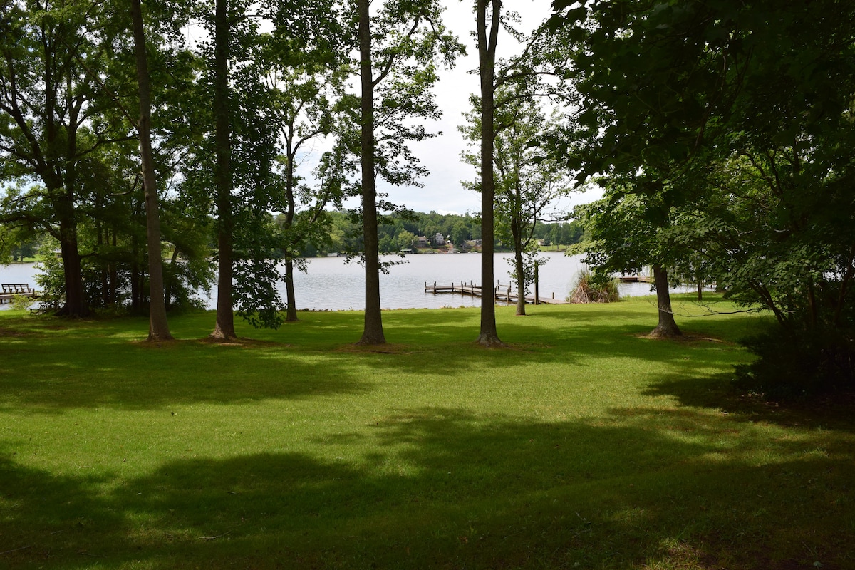A view of a serene lake surrounded by greenery. Tall trees provide shade over a spacious lawn leading to the water's edge, where a dock is visible. The calm lake reflects the sky and surrounding nature, inviting peaceful outdoor activities.