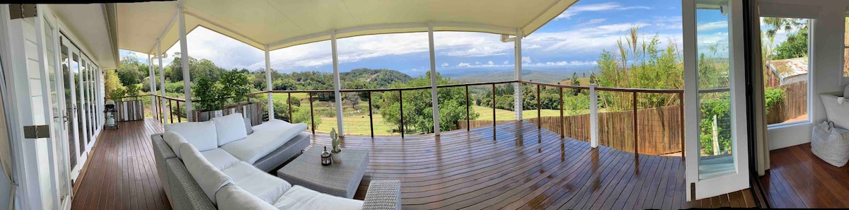 An expansive outdoor deck features comfortable seating and overlooks rolling hills and the lush landscape. Panoramic glass railings enhance the views, while a vibrant sky adds depth to the serene atmosphere. The wooden deck provides a warm contrast to the greenery beyond.