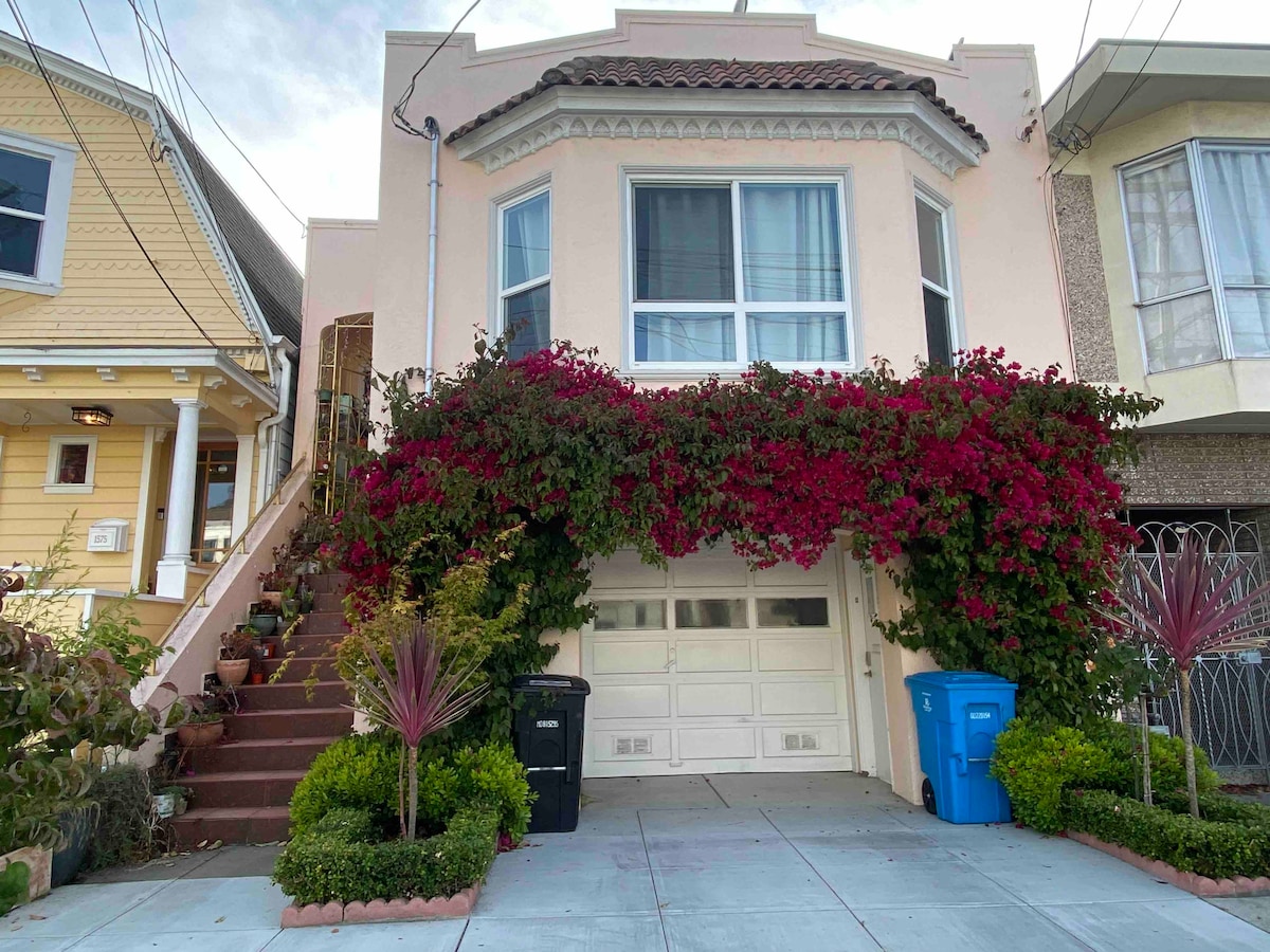 The exterior of the property features a charming facade with a flowering bougainvillea cascading over the entrance. A well-maintained sidewalk leads to the door, with a staircase on one side and vibrant greenery flanking the driveway, which includes a garage.