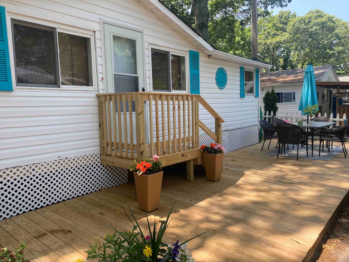 The exterior of the bungalow is presented, showcasing a newly built front deck with a wooden railing. Potted flowers add color to the space. A patio set is arranged on the deck, and a turquoise umbrella provides shade.