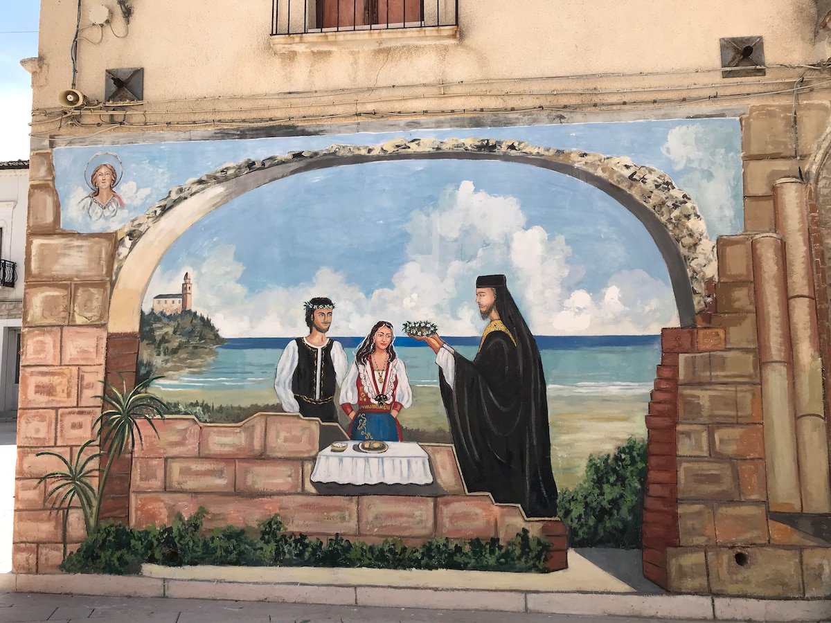 A vibrant mural depicts a scene on the shore, featuring two figures in traditional attire and a cloaked figure officiating a ceremony. An ocean view and building are visible in the background, framed by an archway that adds depth to the outdoor setting.