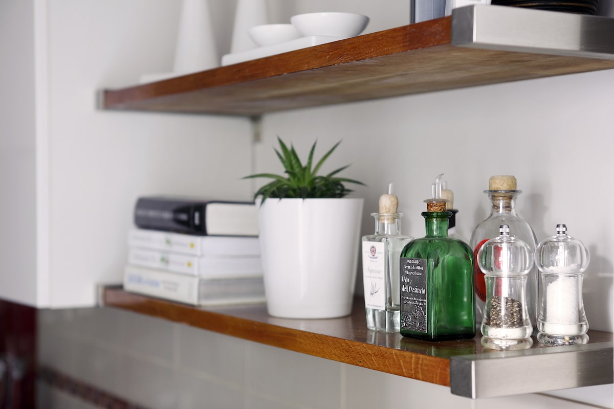 Open shelves display a variety of items, including a small plant in a white pot, kitchen condiments in glass bottles, and several neatly stacked books. Simple dishware is also visible, contributing to a functional kitchen aesthetic.