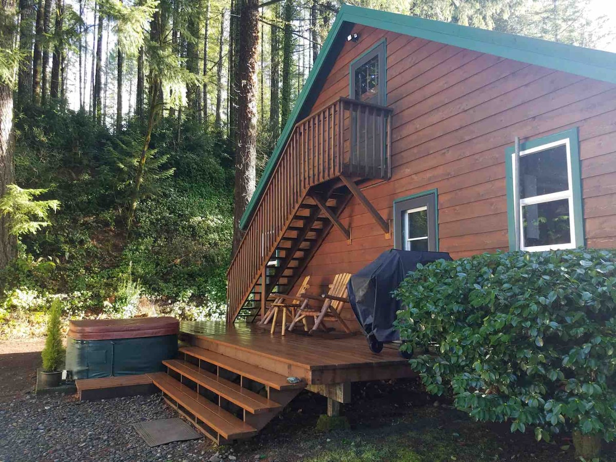 The exterior of the cabin is constructed with rich wood panels and features a green roof. An outdoor deck extends towards the front, equipped with a hot tub and two wooden chairs. An exterior stairwell leads to the upper level, surrounded by lush greenery.