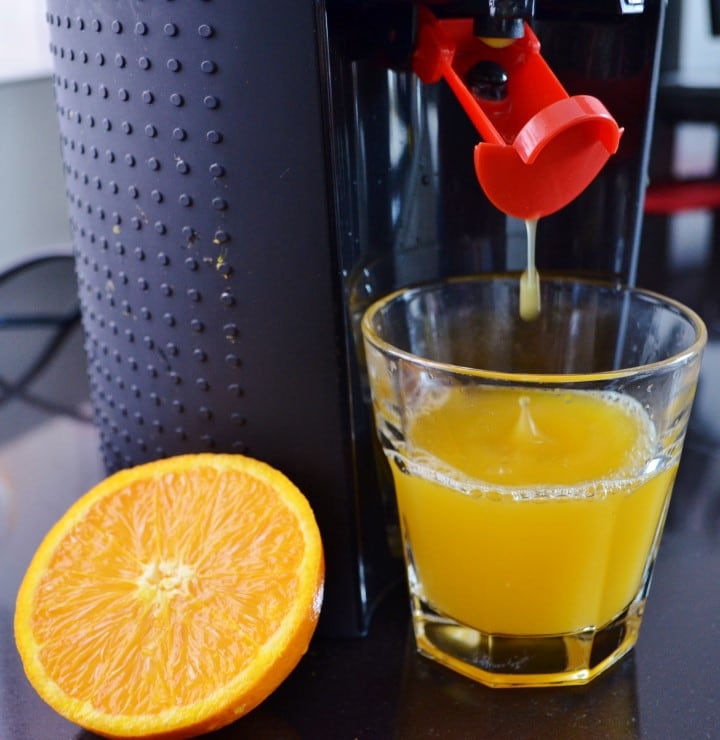 A juicer is seen pouring fresh orange juice into a clear glass. A halved orange rests on the countertop beside the glass, showcasing its vibrant color and texture.