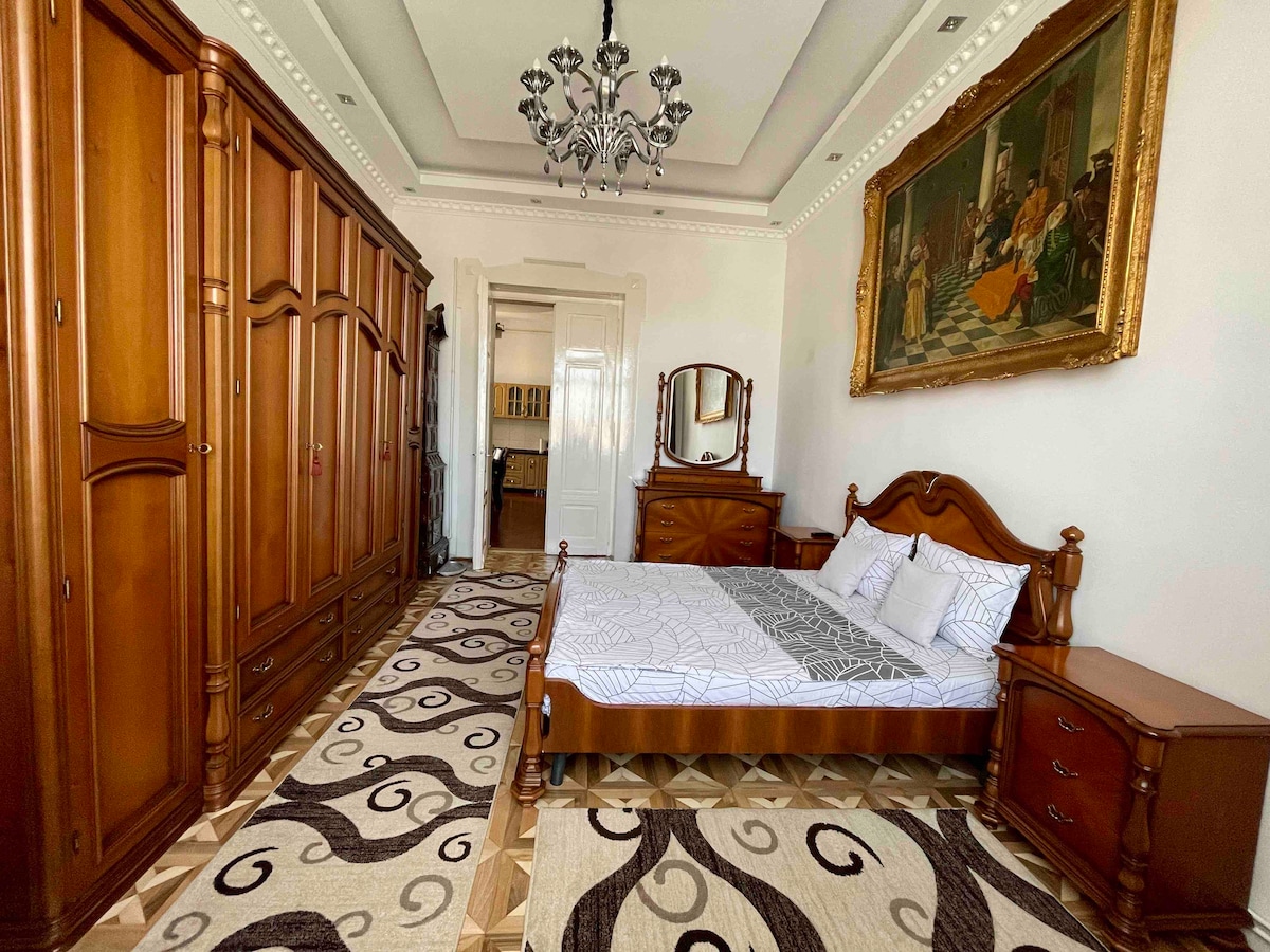 A spacious bedroom features a wooden bed with a patterned coverlet. A large wardrobe and matching nightstands provide storage options. An ornate chandelier hangs from the ceiling. A decorative area rug complements the room's neutral tones, while a painting adorns the wall above the bed.