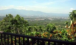 A panoramic view of the surrounding landscape is visible from the balcony. Lush greenery and vibrant flowers frame the scene, which includes distant mountains under a clear blue sky. The expansive vista offers insight into the natural beauty of the area.
