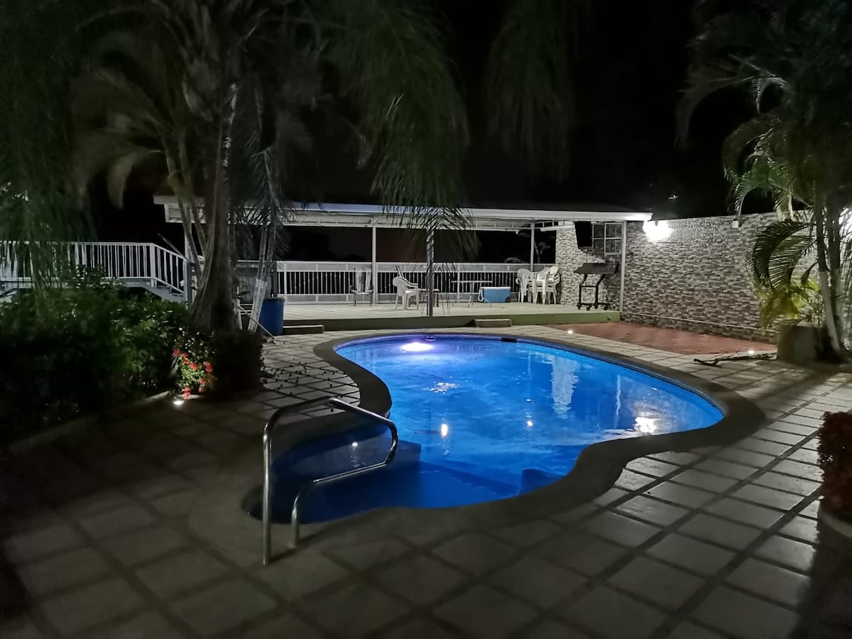 A swimming pool is illuminated at night, creating a calming blue glow. The surrounding area features tiled flooring and lush palm trees, with a covered outdoor space visible in the background, complete with seating. Soft lighting enhances the serene atmosphere.