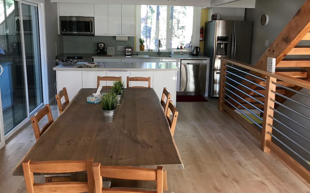 A spacious kitchen and dining area is highlighted by a large wooden table surrounded by six chairs. Light-colored hardwood floors extend throughout the room, while large windows provide natural light. The kitchen features modern appliances and clean white cabinetry, creating an open and airy atmosphere.