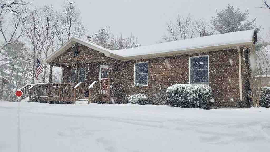A charming cabin is shown amidst falling snow, covered in a soft, white blanket. The entrance features a wooden porch with railings and an American flag, surrounded by snow-laden shrubs. The landscape emphasizes a serene winter setting.