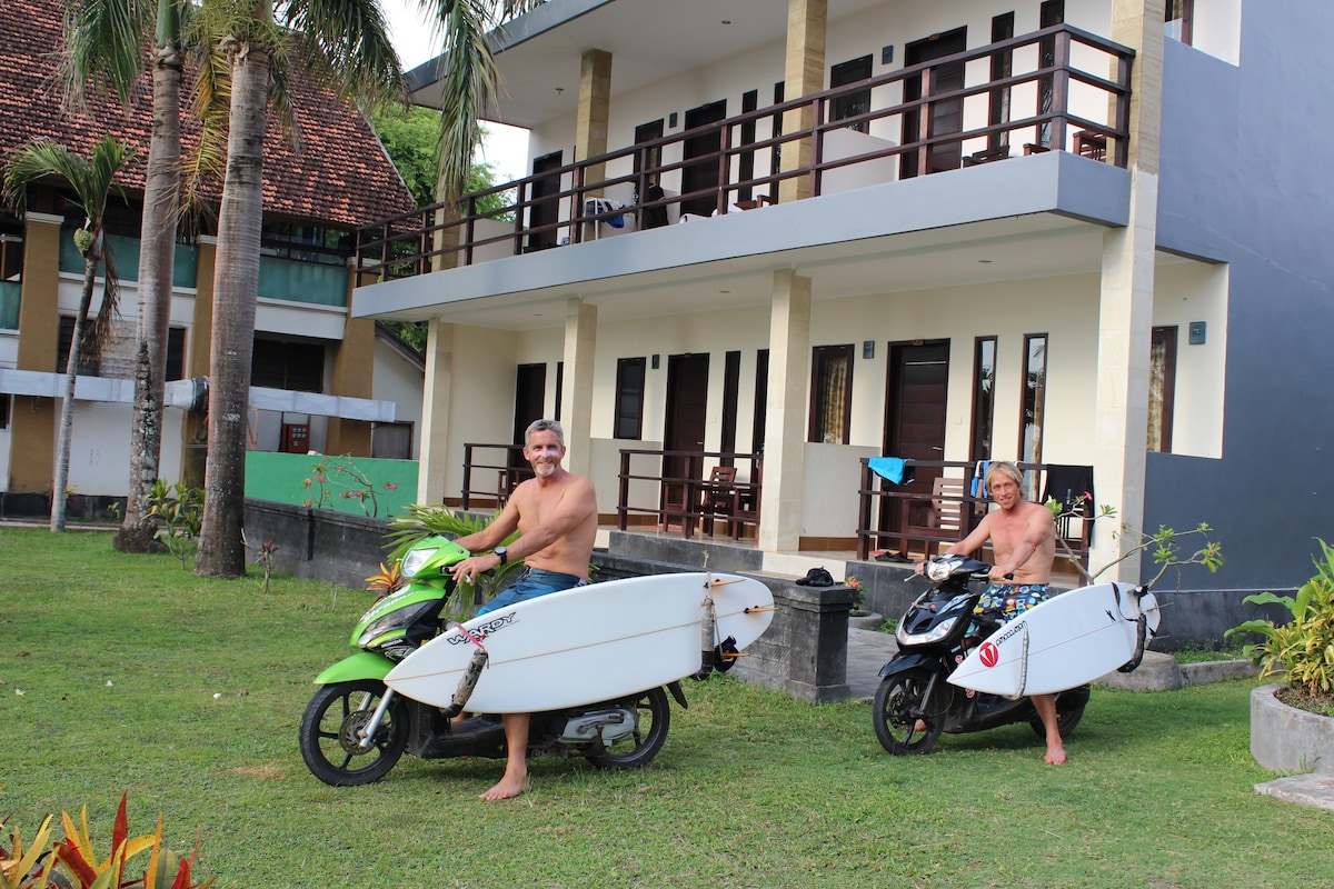Two individuals are seen preparing for surfing, with surfboards mounted on scooters. The background features a multi-story building with balconies and several doors, surrounded by lush green grass and tropical plants, creating a casual outdoor atmosphere.