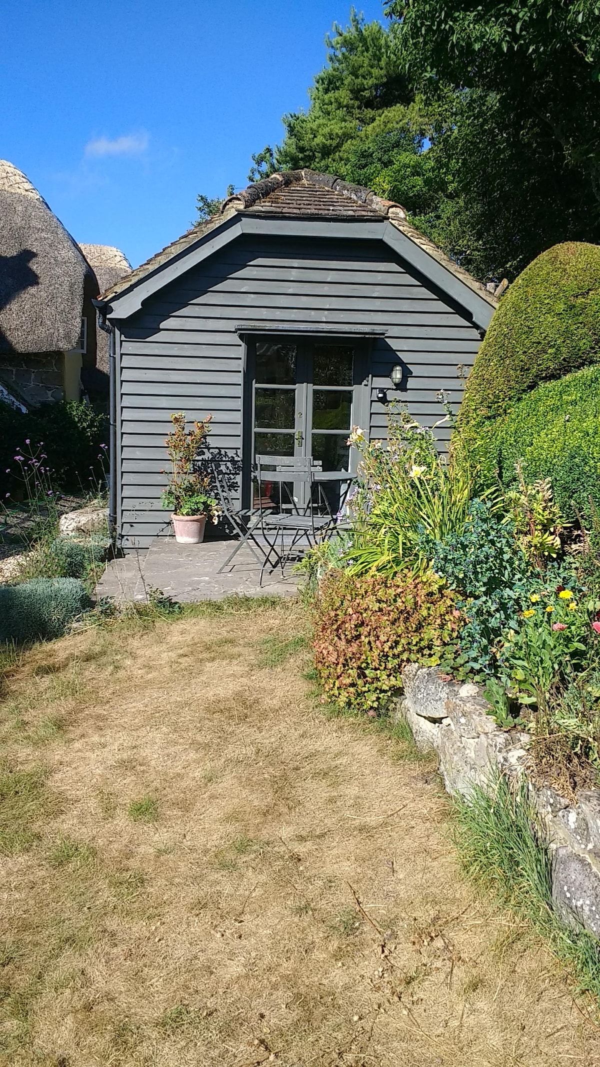 A charming garden room features a gray exterior and a door framed by large glass windows. A small outdoor terrace is set with a table and chairs, surrounded by lush greenery and blooming flowers, creating an inviting outdoor space.